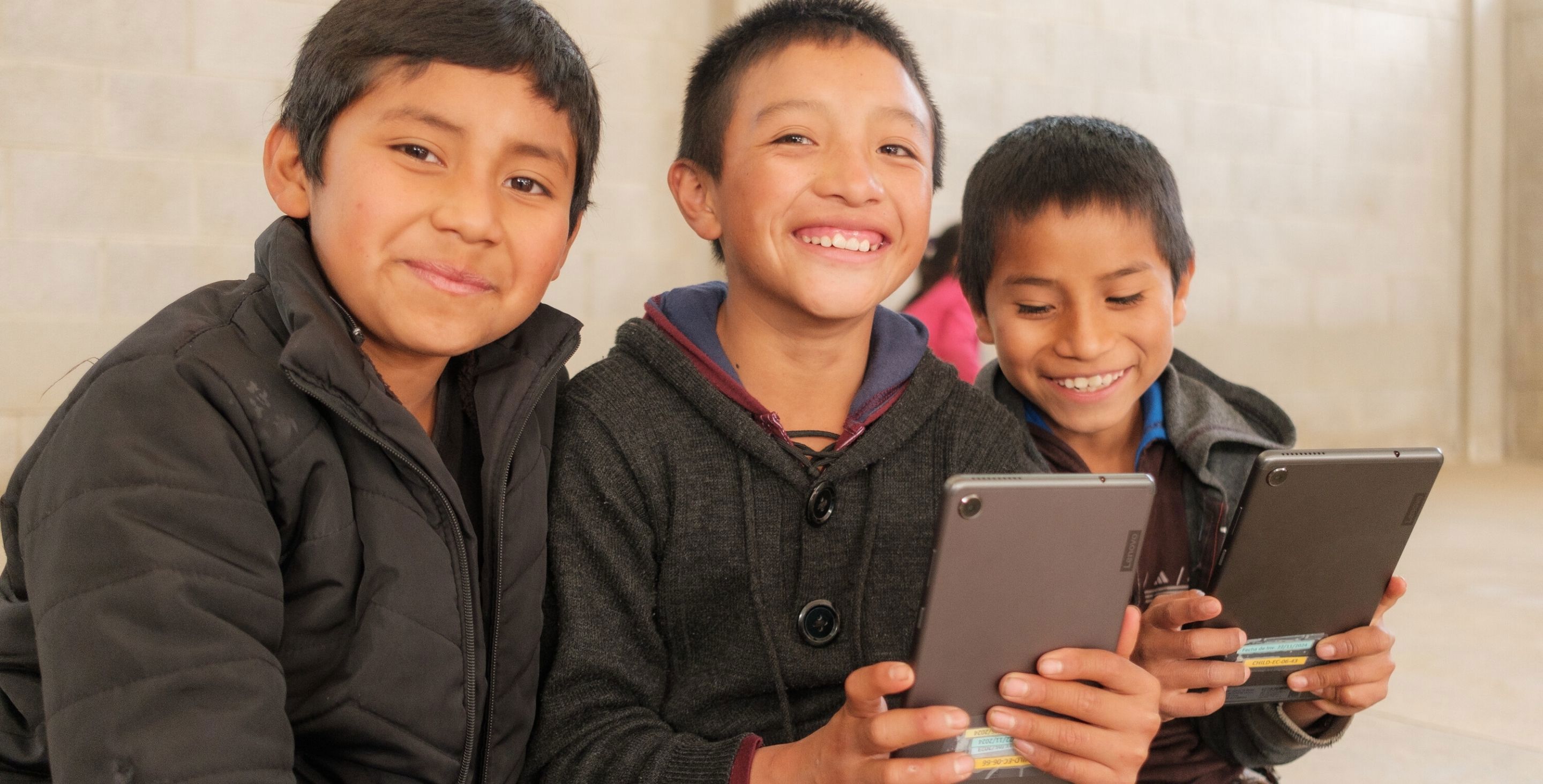 Three boys in Guatemala sit at a desk while looking at cell phones together and smiling.