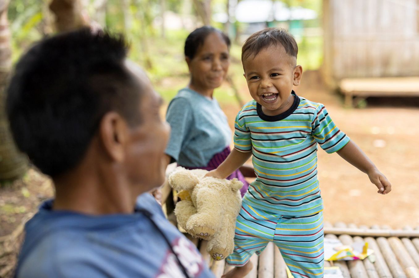 A toddler in a striped outfit laughs while walking toward his father on a bamboo platform in Indonesia.