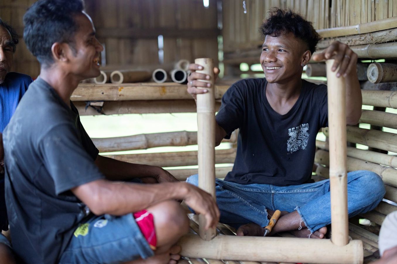 A young man in his 20s laughs while sitting in a bamboo home in Indonesia. He is holding materials for woodworking and talking to his instructor, who is seated beside him.