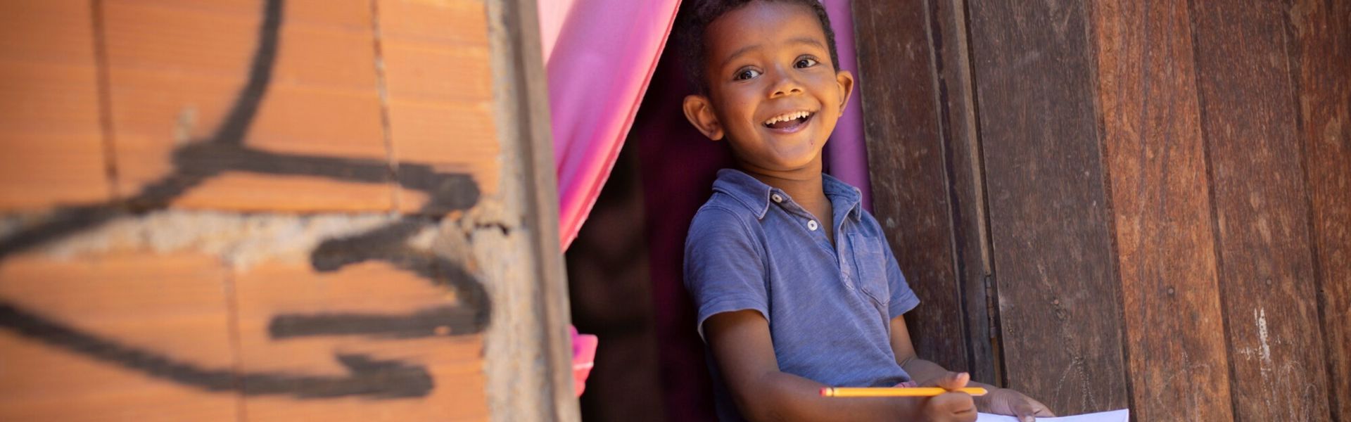 A child in Brazil sits in a doorway, holding a pencil and piece of paper in hand and smiling at the camera.