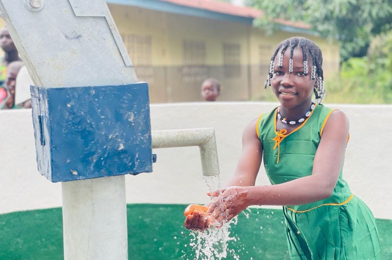 A teenage girl in Sierra Leone with her hair in braids washes her hands at an outdoor water faucet.