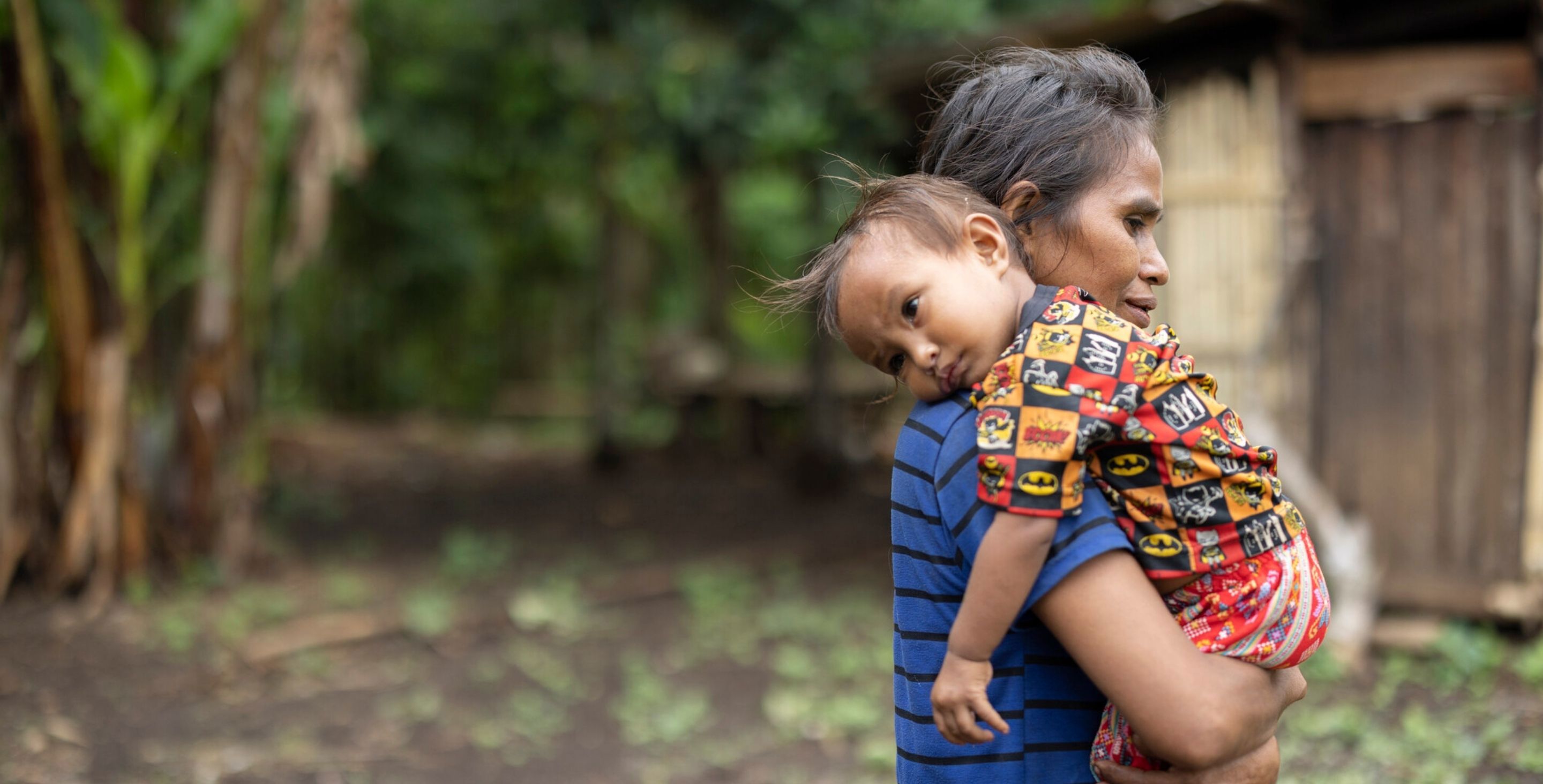 A mother in Indonesia carries her toddler over her shoulder. The toddler has a sad, somber expression on her face.