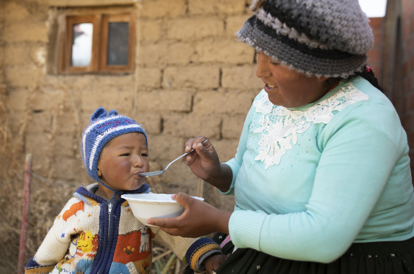 A mother in Bolivia feeds her toddler boy from a bowl while sitting in front of their home.
