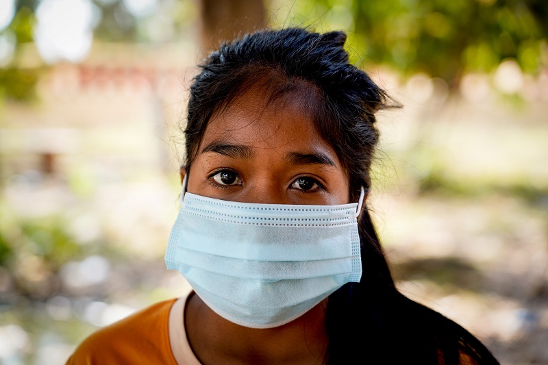 A young teenage girl wearing a mask in Cambodia looks at the camera pensively.