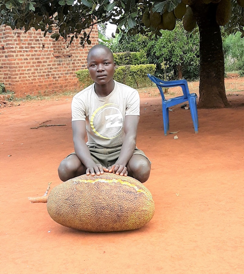 A young man sits on the ground in Uganda behind a giant jackfruit, looking solemnly at the camera.