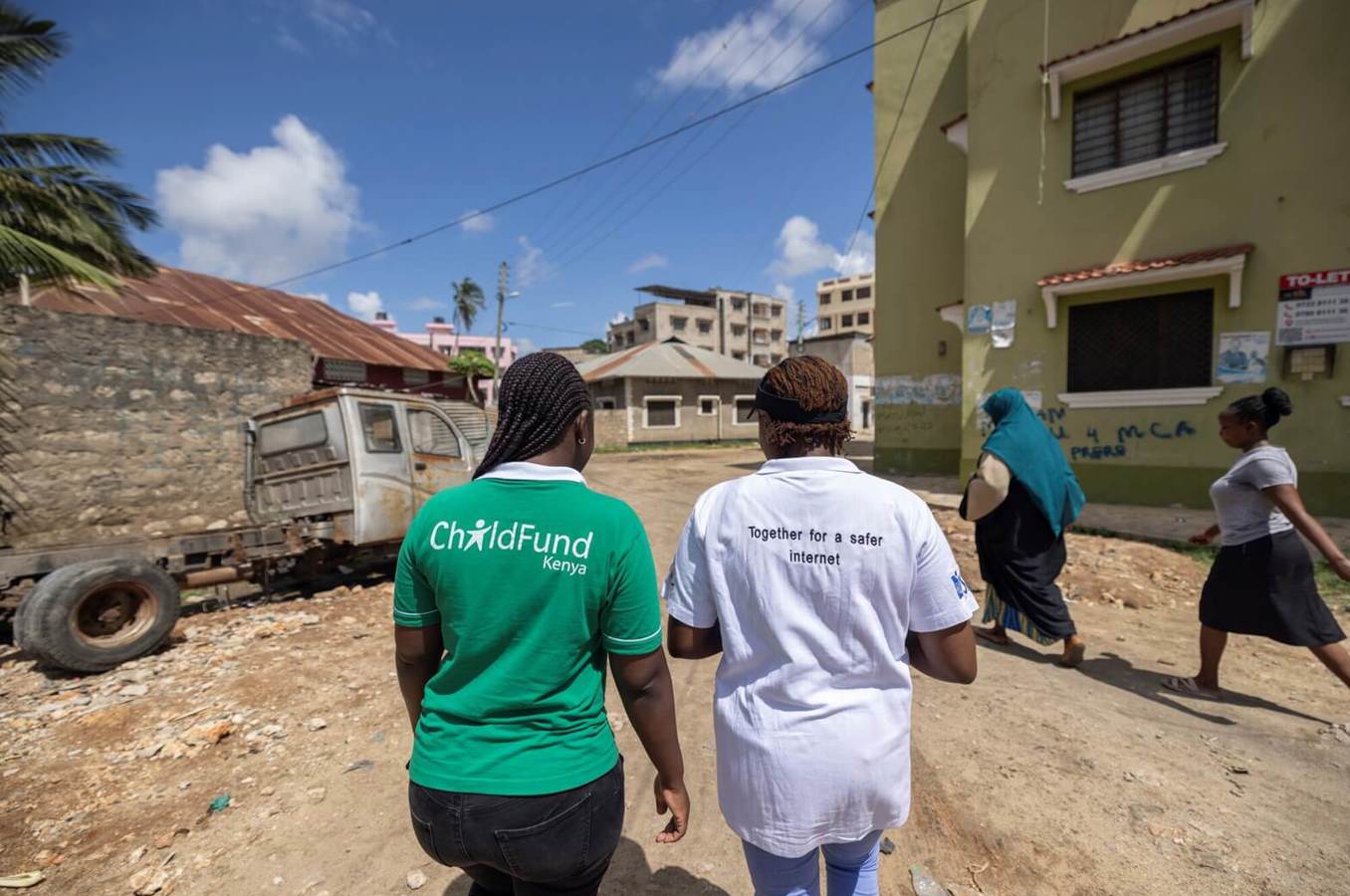 Two people wearing ChildFund Kenya and online safety project shirts walk through a residential area, with other community members and buildings visible around them.