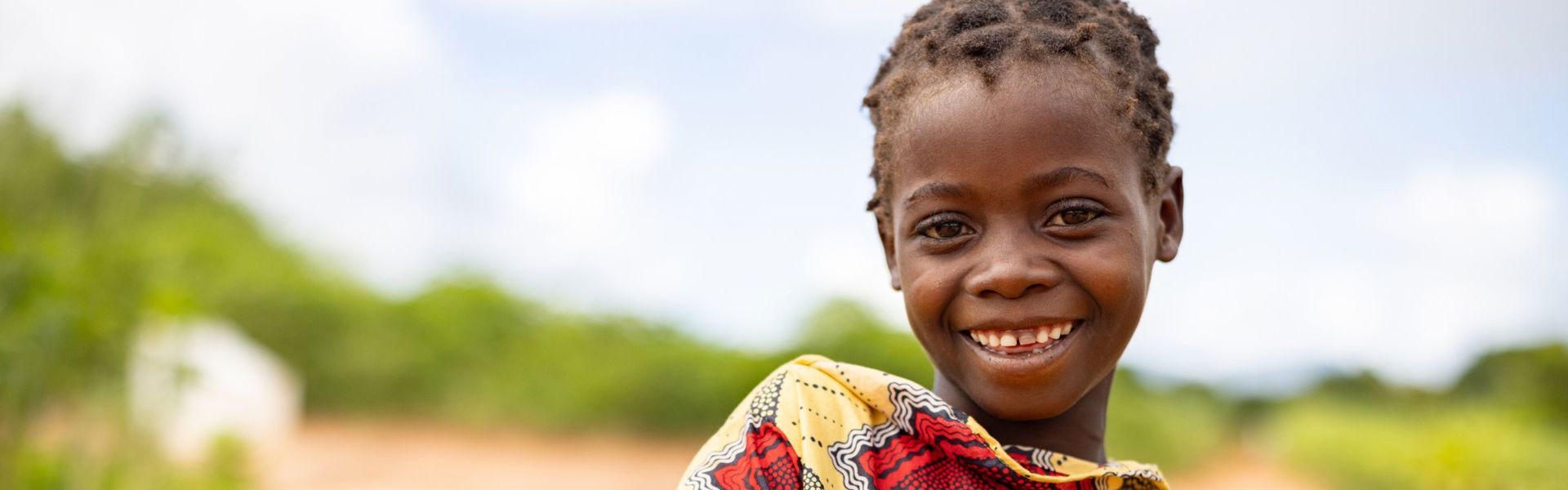 Little girl in Mozambique wearing traditional dress stands in a dirt road, smiling.