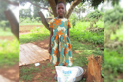 A young girl in Uganda stands outside with bag of flour, food, pots and pans. 