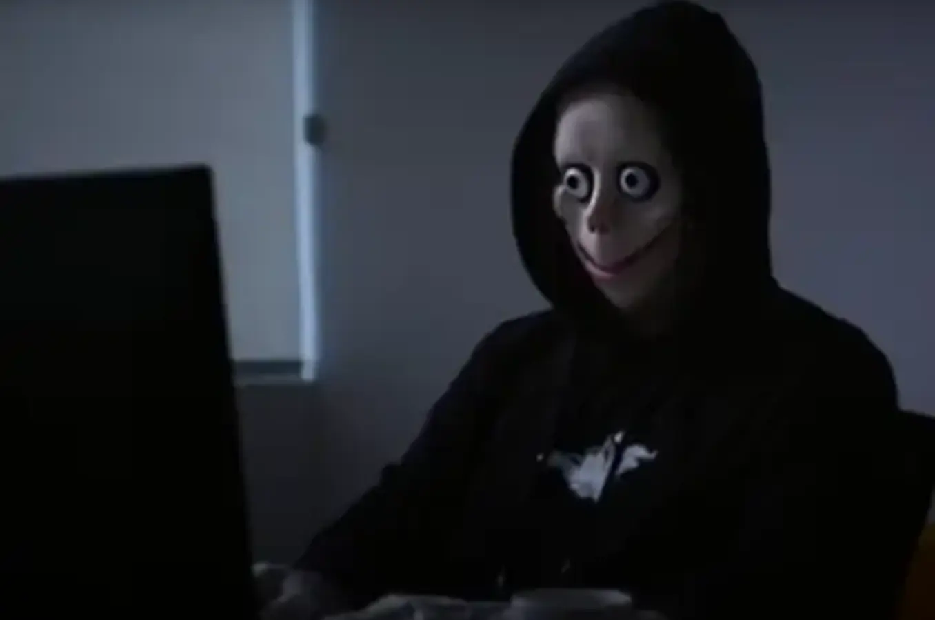 A hooded figure sits in a dark room using a laptop, symbolizing online threats and the need for internet safety awareness.