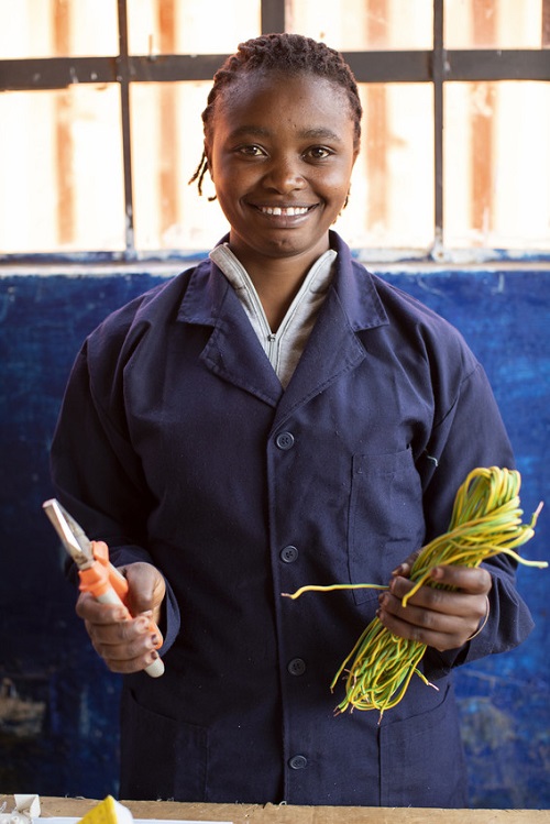 Girl in Kenya stands holding electrical tools and looking at camera smiling.