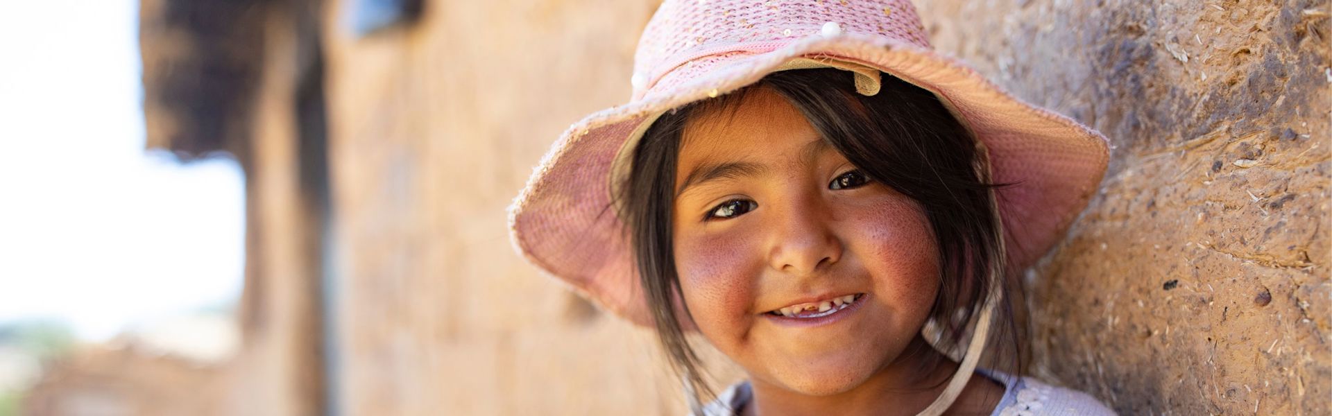 A girl in rural Bolivia wearing a pink hat stands outside of her home, smiling.