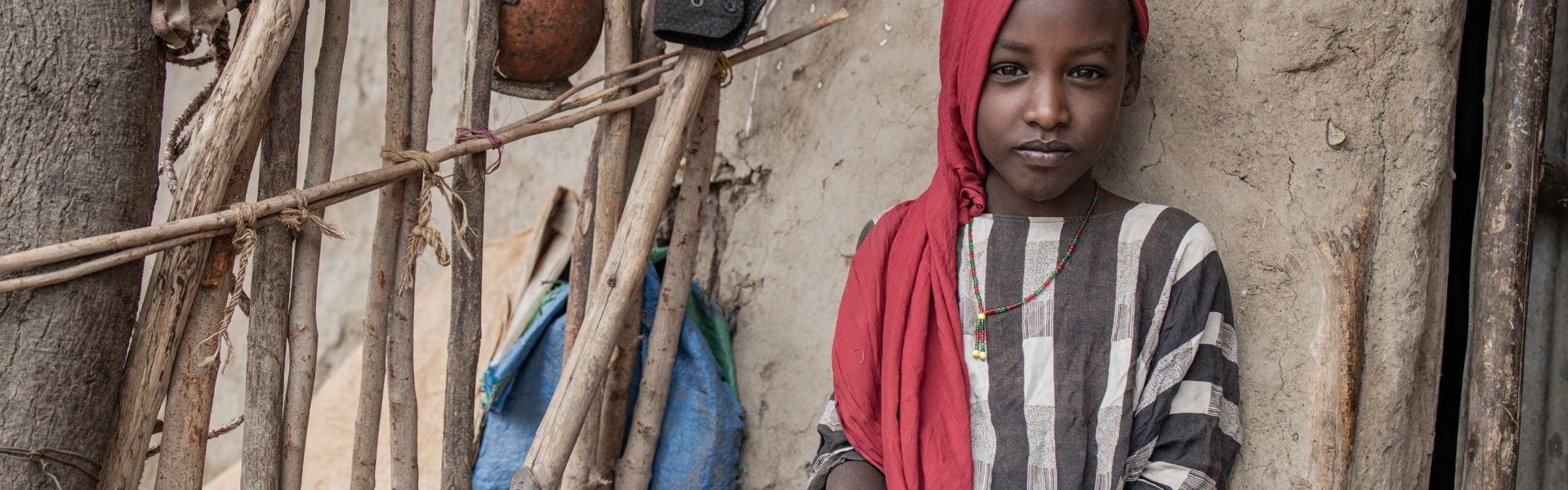 A young girl in Ethiopia wearing a red head scarf sits against the outer wall of a hut, preparing maize in a basket. She is looking somberly at the camera.