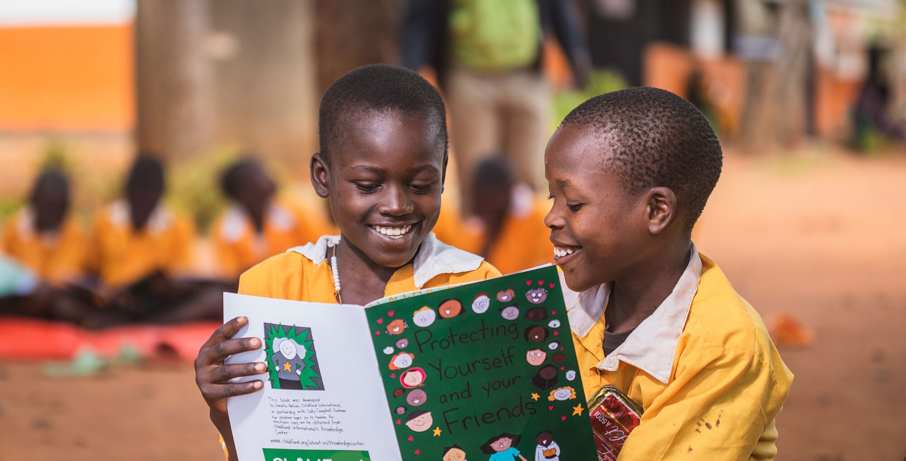 Two little boys in Uganda sit on the ground reading a book called 