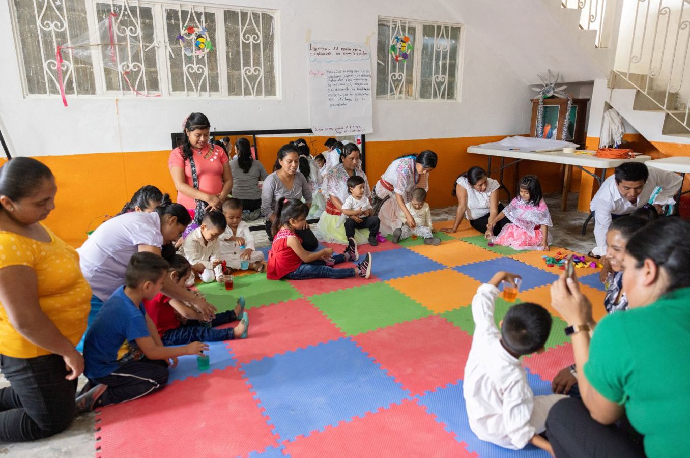 A group of parents and toddlers in Mexico gather around a large colorful rug, smiling and playing.