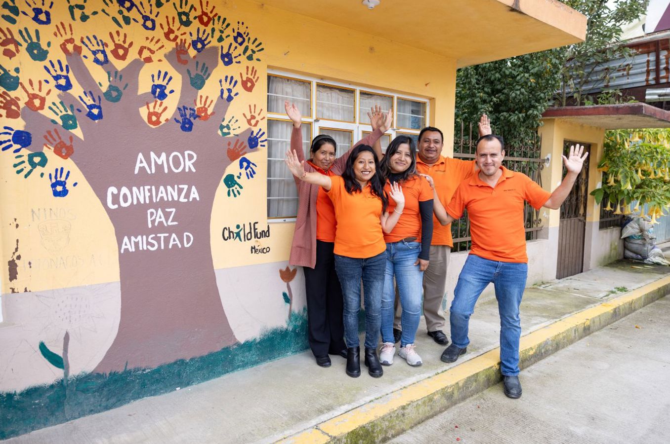 A group of men and women in orange shirts stand outside an office building in Mexico. There is a colorful mural behind them featuring a tree with different words on it in Spanish.