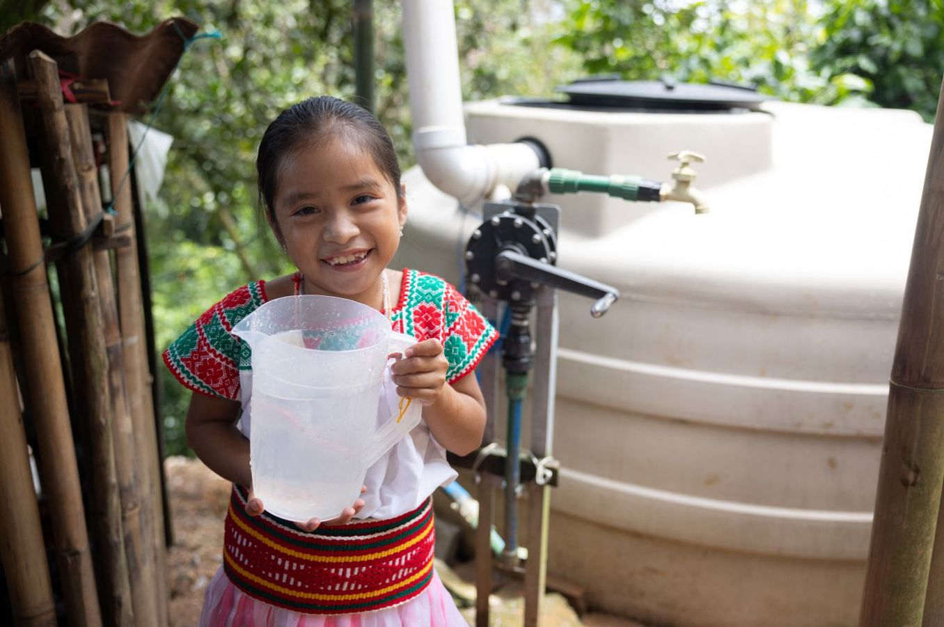 A little girl in Mexico wearing traditional clothing stands in front of a large water tank, holding a pitcher of clean water and smiling.