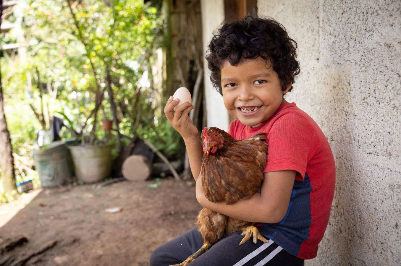 A little boy in Mexico smiles while sitting on his doorstep, holding a chicken under his arm and an egg in the other hand.