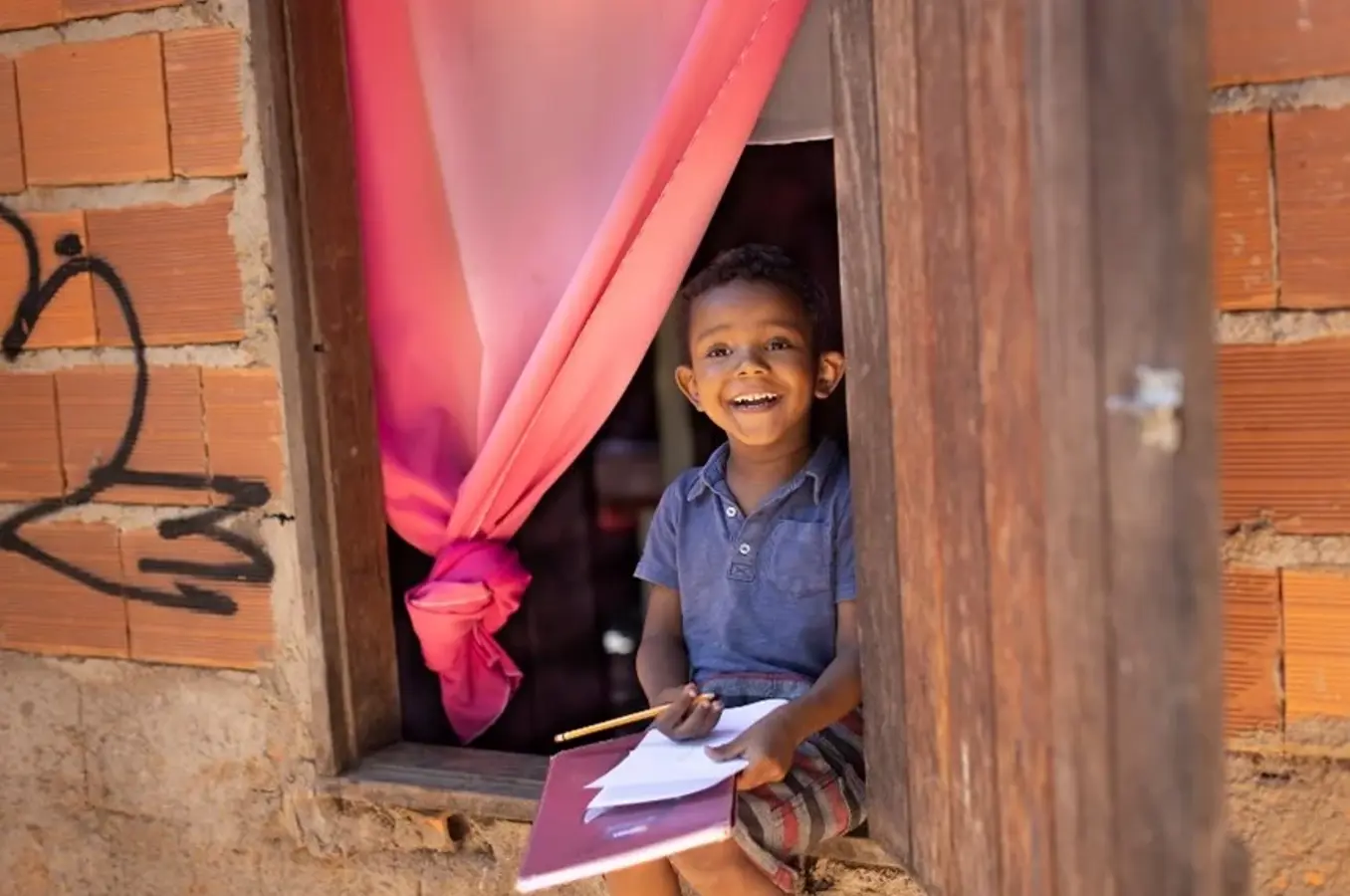 A young boy smiles while drawing in a notebook.