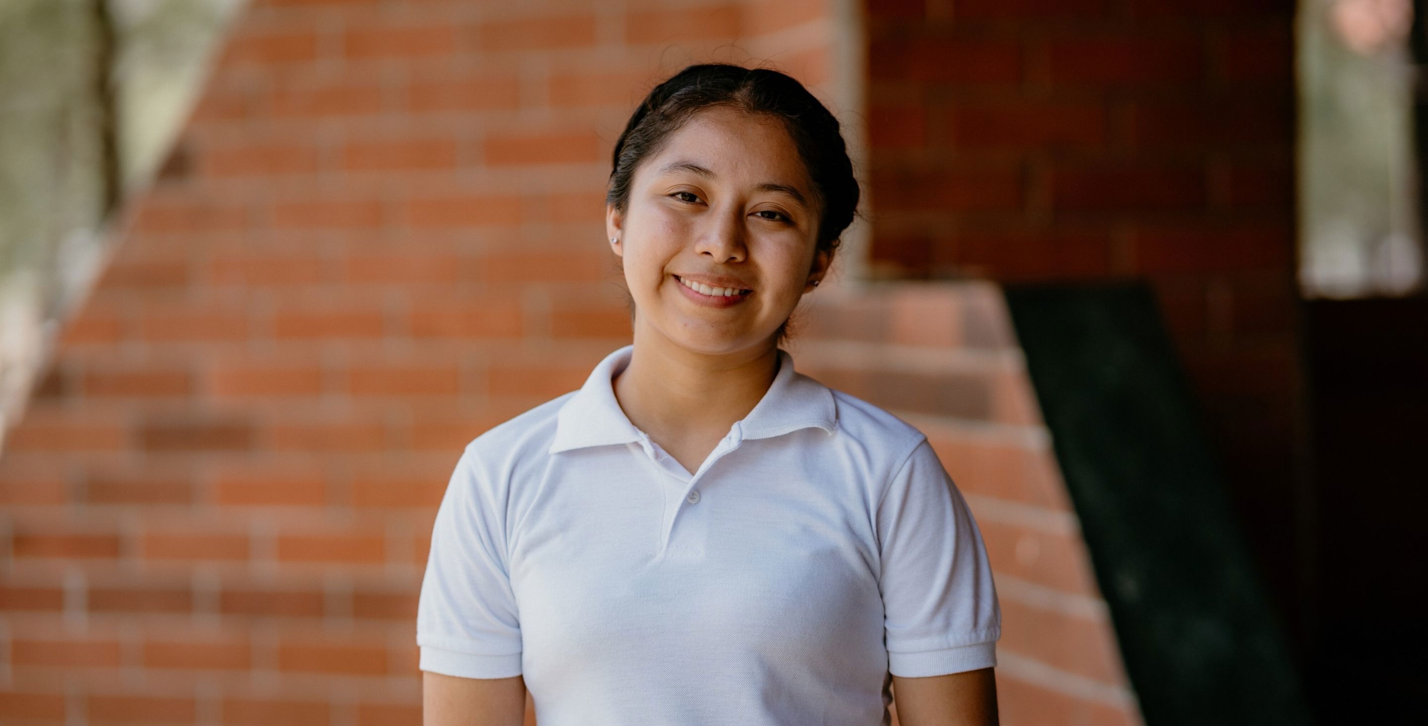 A young woman in Guatemala wearing a white shirt stands against a brick wall, smiling at the camera.