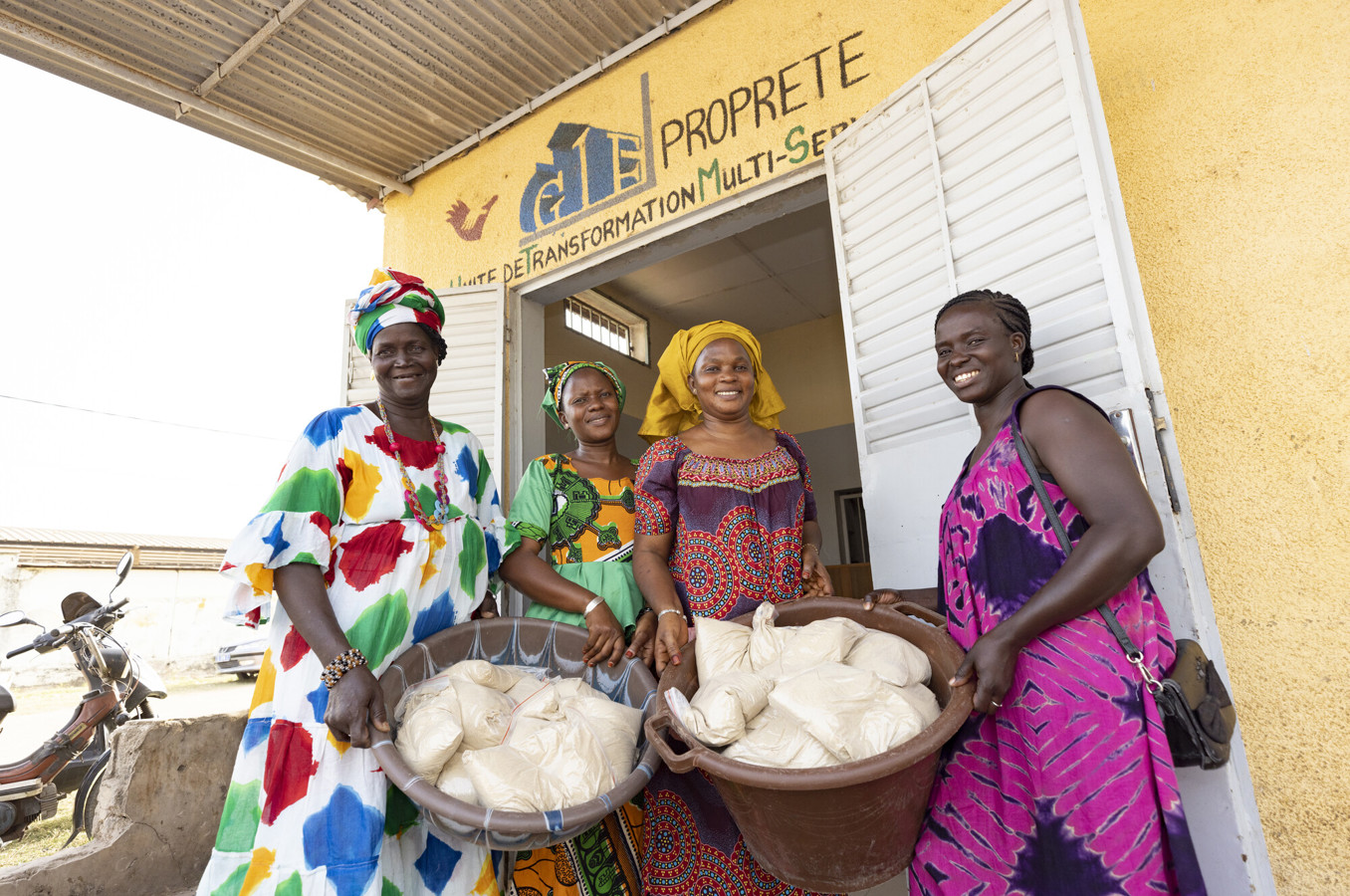 A group of colorfully dressed women in Senegal stand in front of a doorway, holding large baskets with bags of nutritional porridge.