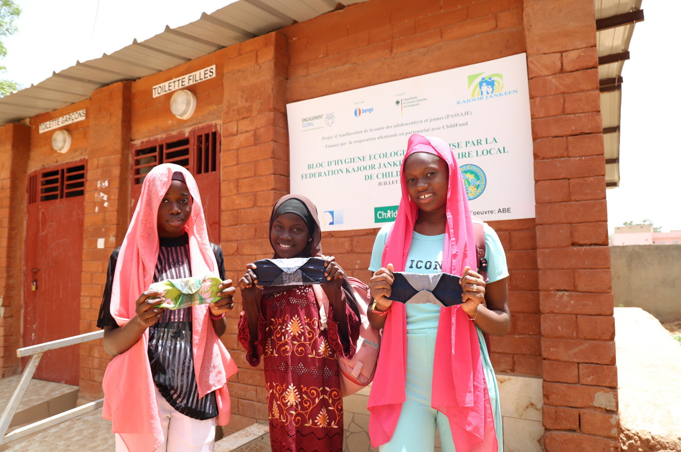 Three teen girls in Senegal stand in front of a washroom, holding homemade sanitary pads.