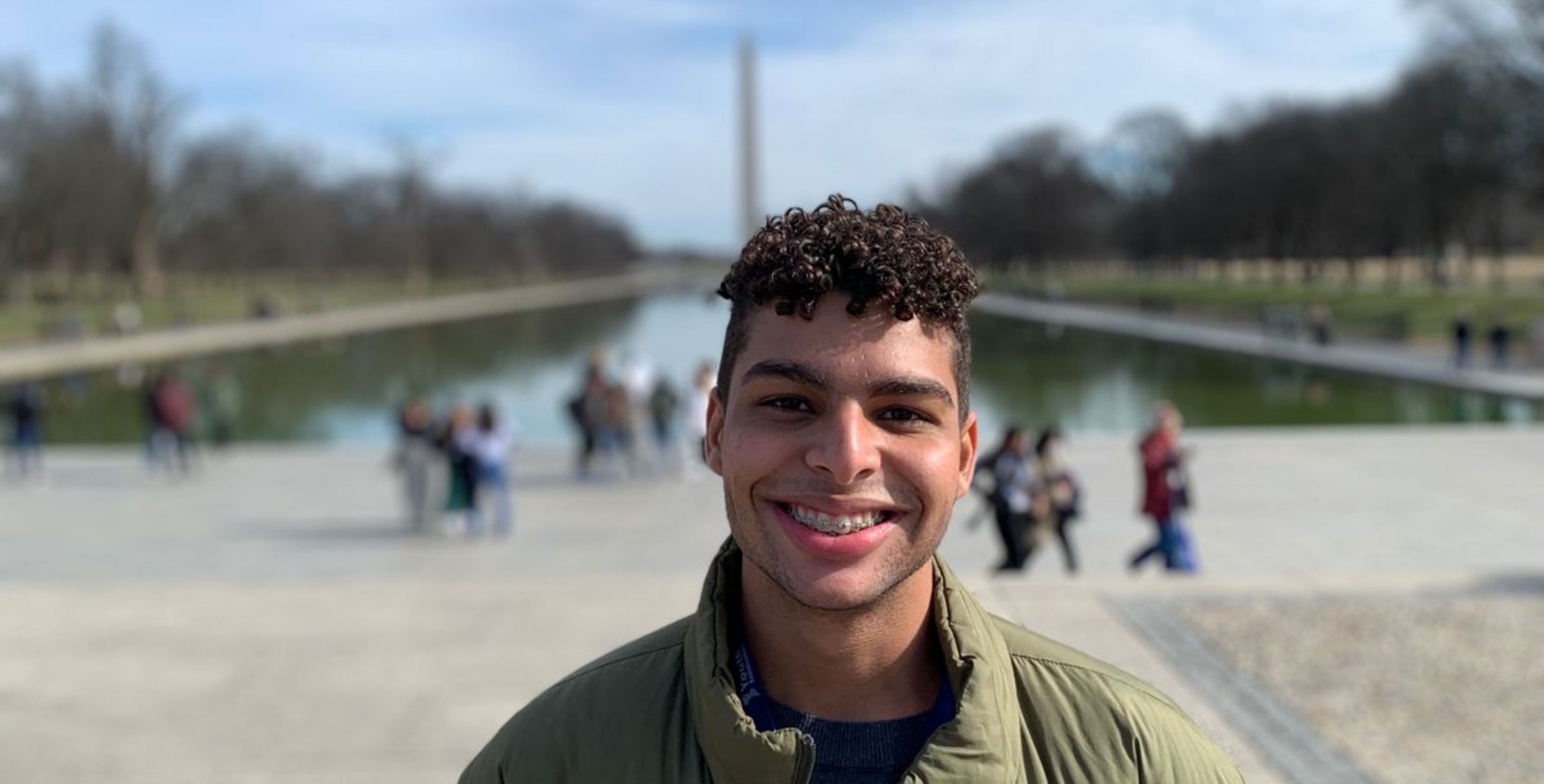 A young man from Brazil stands in front of the Washington Monument in the U.S., smiling widely.