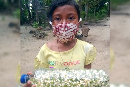 Girl wearing facemask stands outdoors holding bottle filled with sprouts in Indonesia.