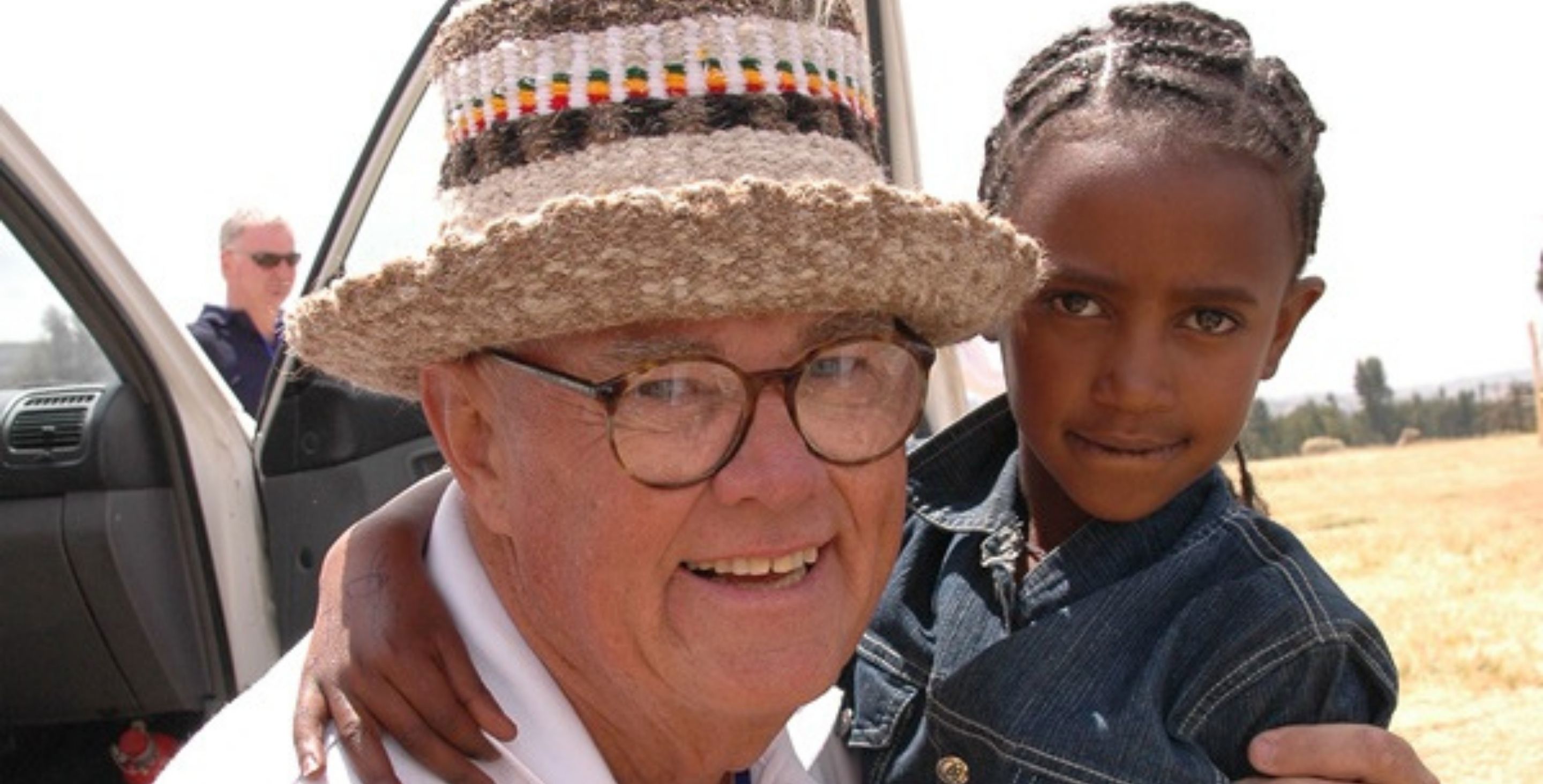 An American man in a straw hat and a young girl in Ethiopia pause for a photo together in a field.