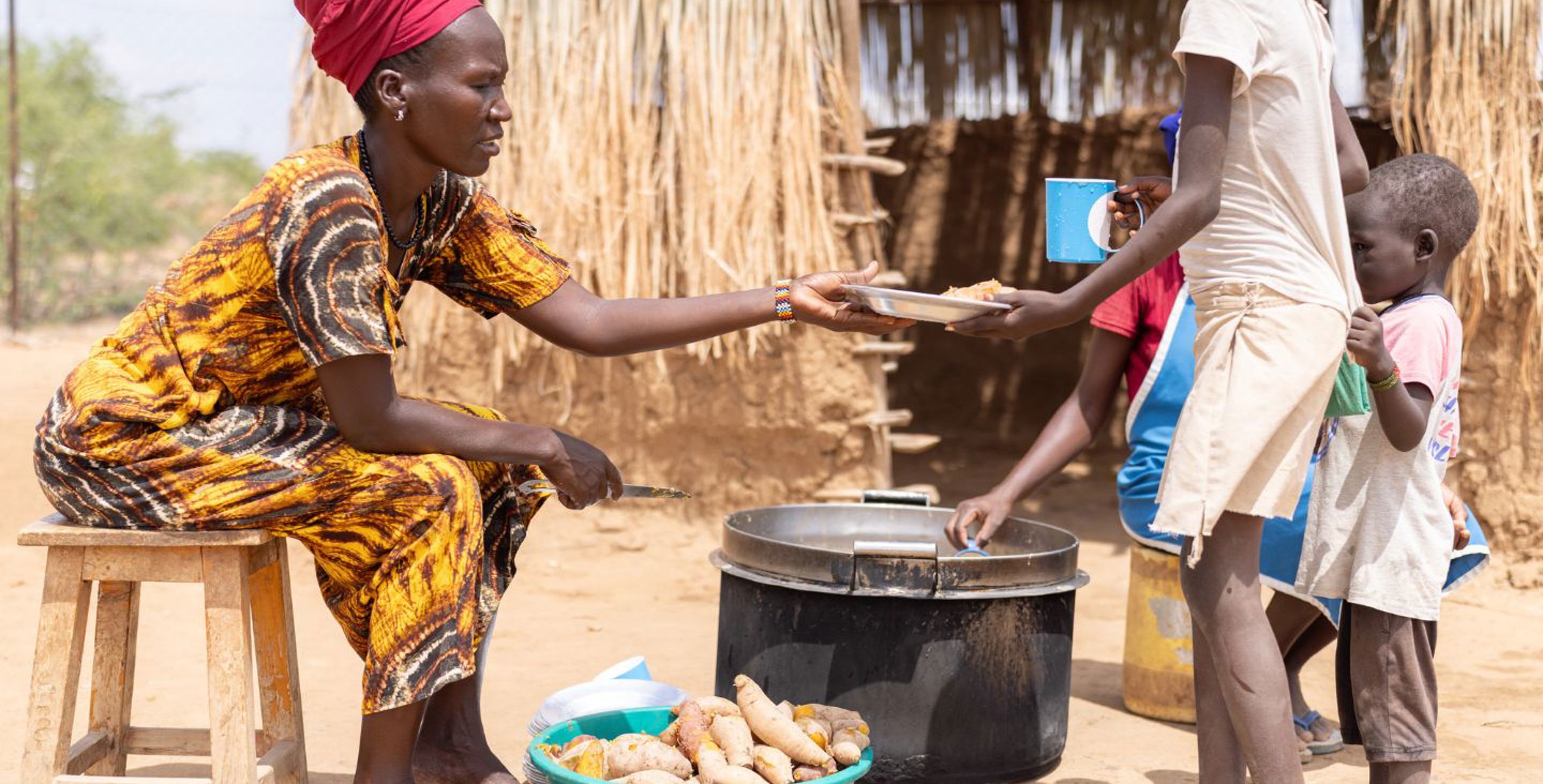 A woman wearing a traditional dress and head wrap in Kenya serves plates of sweet potatoes to children standing in a line, serving the food from a large pot at which she sits.