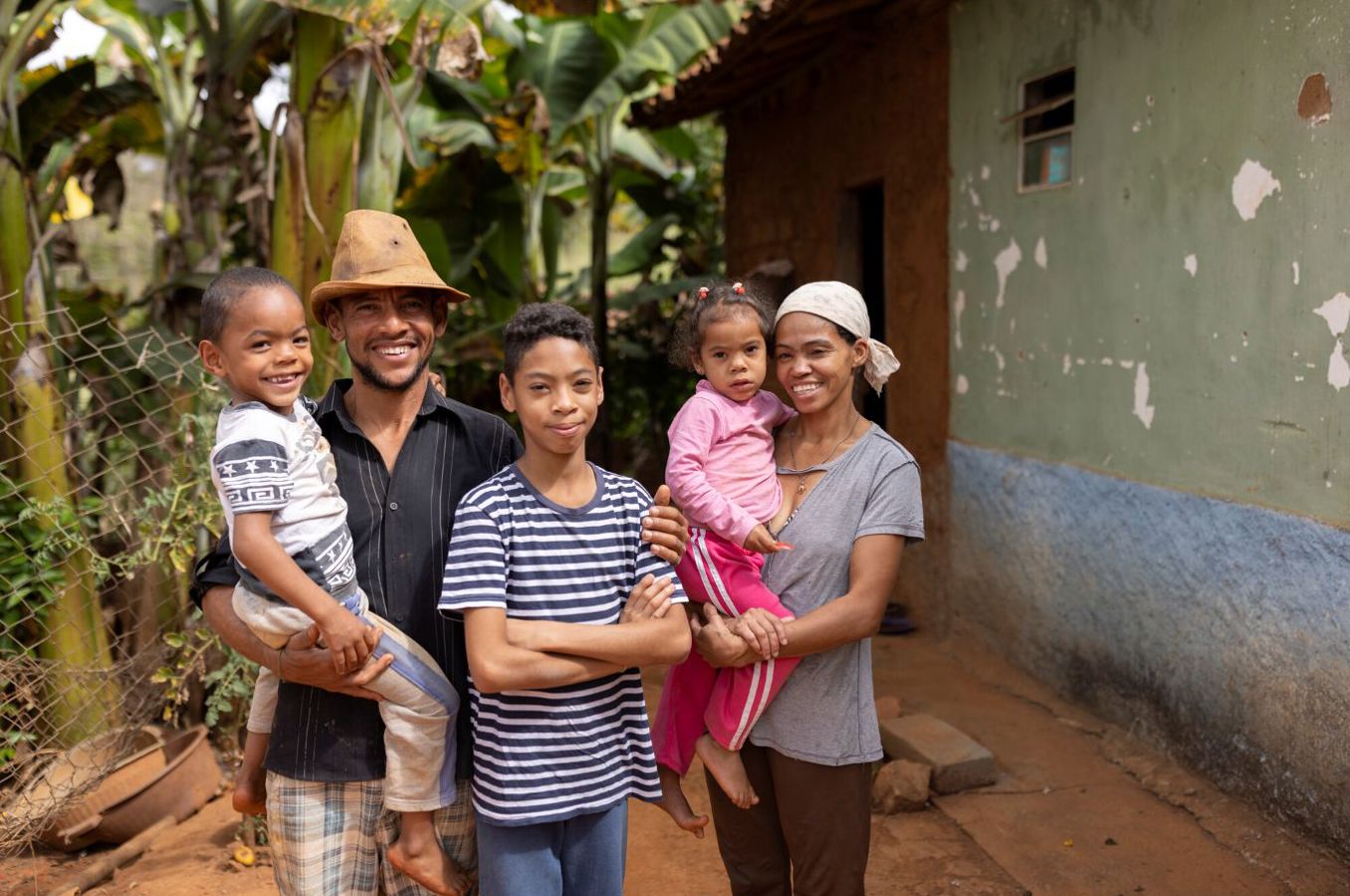 A family consisting of a mom, dad and three children stand in front of their home in Brazil, smiling. The mom is holding her preschool age daughter, the dad is holding his preschool age son, and an older boy stands in the middle.