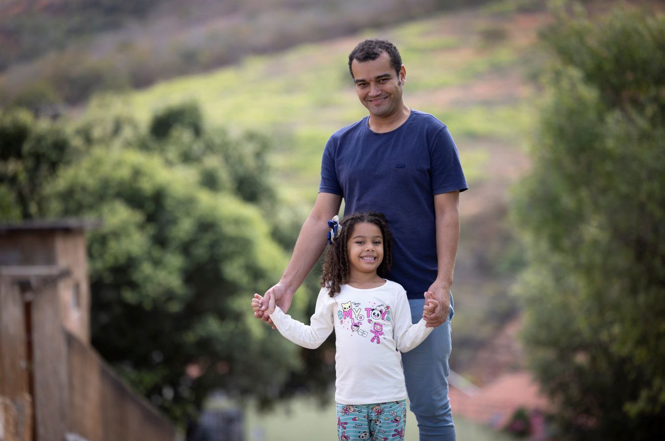 A man stands behind his daughter on a road, holding her hands. Both are smiling at the camera.