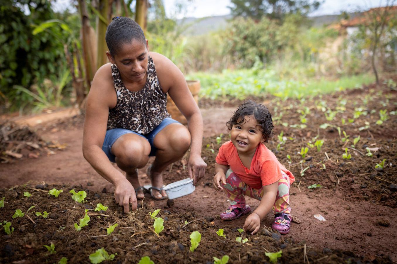 A mom and 2-year-old girl work in the garden together, the girl smiling at the camera.