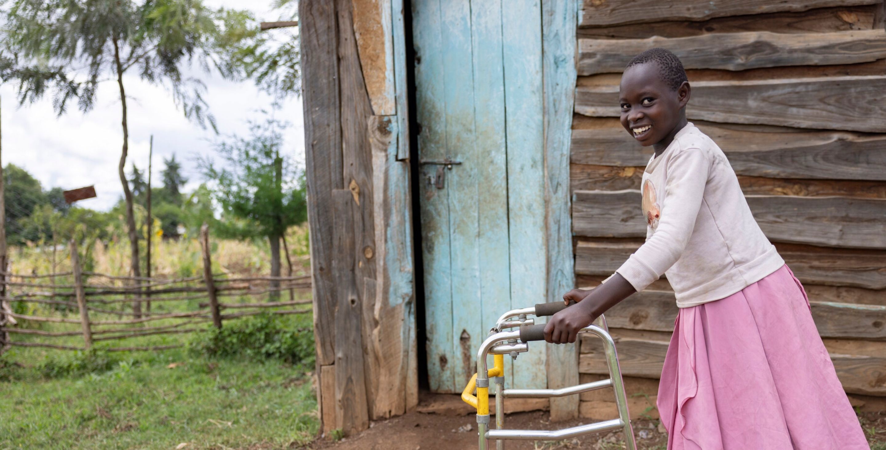 A young girl in Kenya wearing a dress and a sweater walks in front of her hut with a walker.