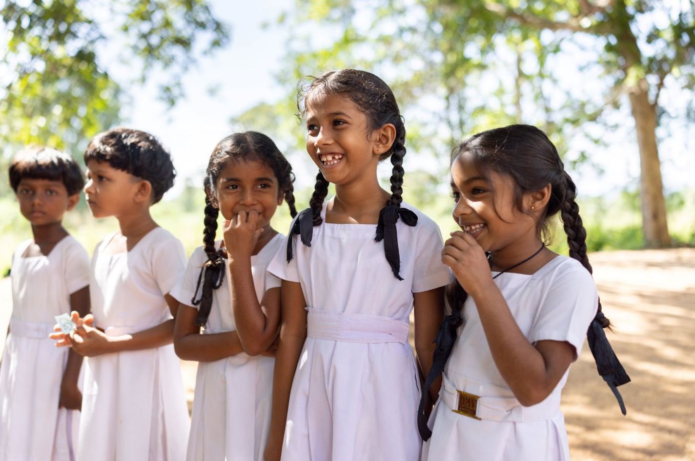 A group of girls in white dresses and pigtails stand in a line, laughing, outdoors in Sri Lanka.