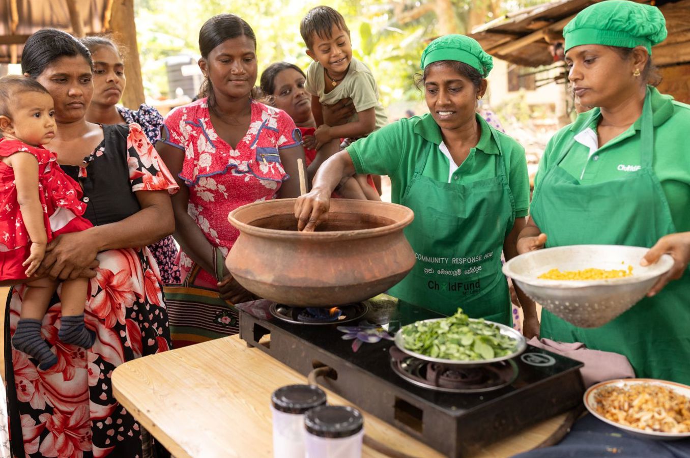 A group of women in hair nets and ChildFund shirts serve food from enormous pots to mothers and children.