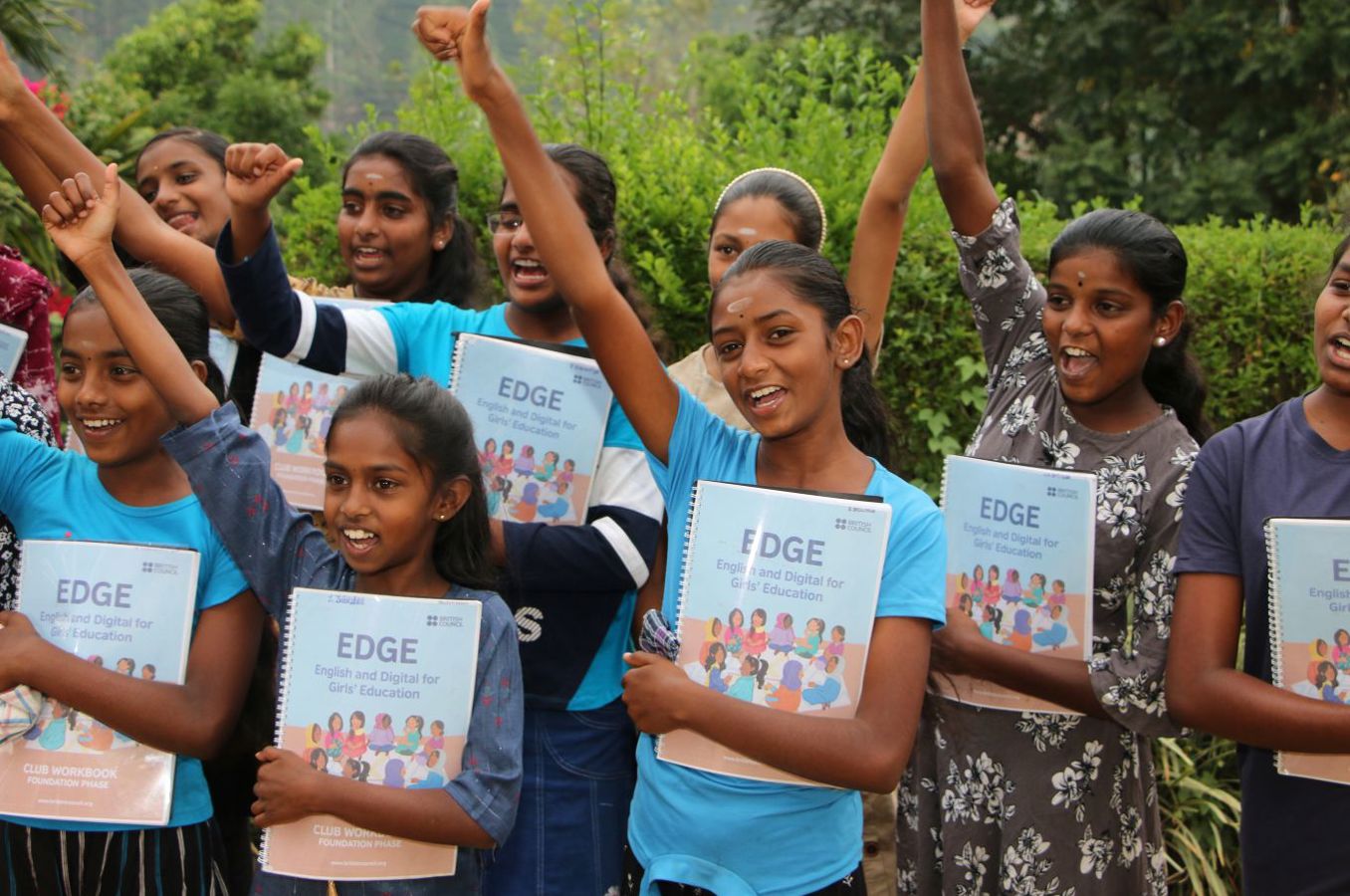 A group of teenage girls wearing school uniforms and clutching notebooks raise their hands in a cheer.