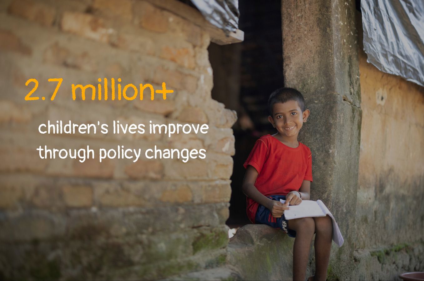 Photo of a boy sitting in the doorway of a mud house, studying. Text on photo reads 
