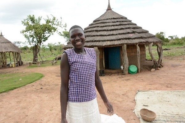 Young girl smiles at the camera in Uganda, a hut behind her.