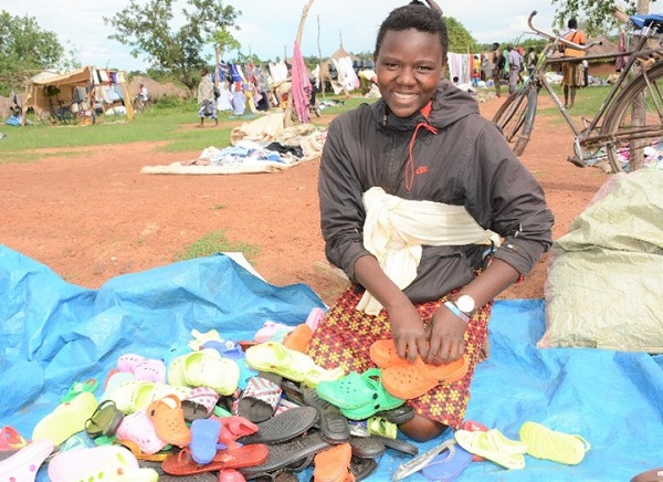 Girl sits on blanket at market in Uganda surrounded by shoes and smiling.