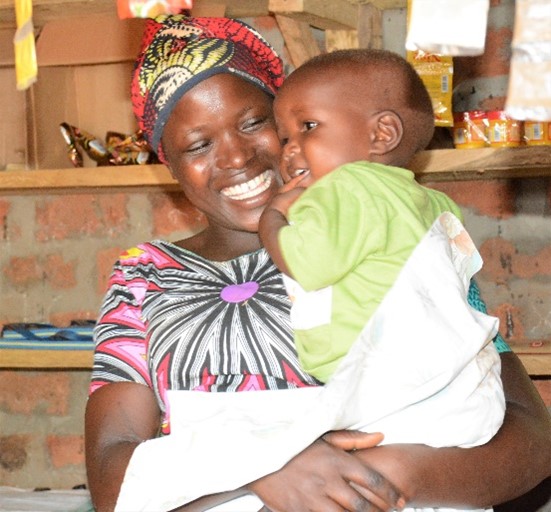 A young woman in Uganda holds a baby in a grocery shop, smiling.