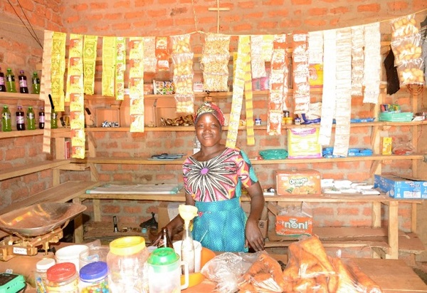 A woman in Uganda stands inside her grocery shop, smiling at the camera.