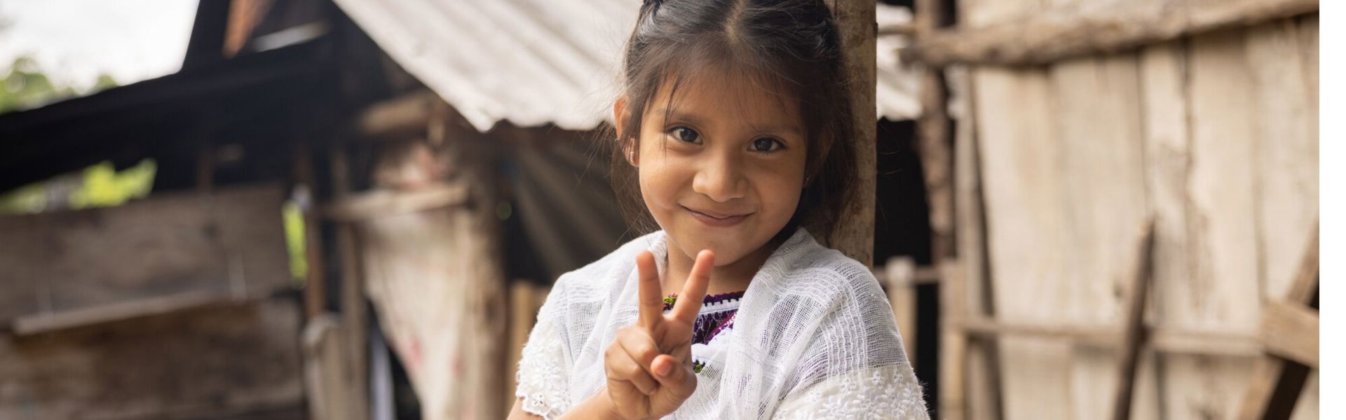 A young girl in Mexico wearing a traditional dress flashes a peace sign at the camera, smiling slightly, outside her home in rural Mexico.