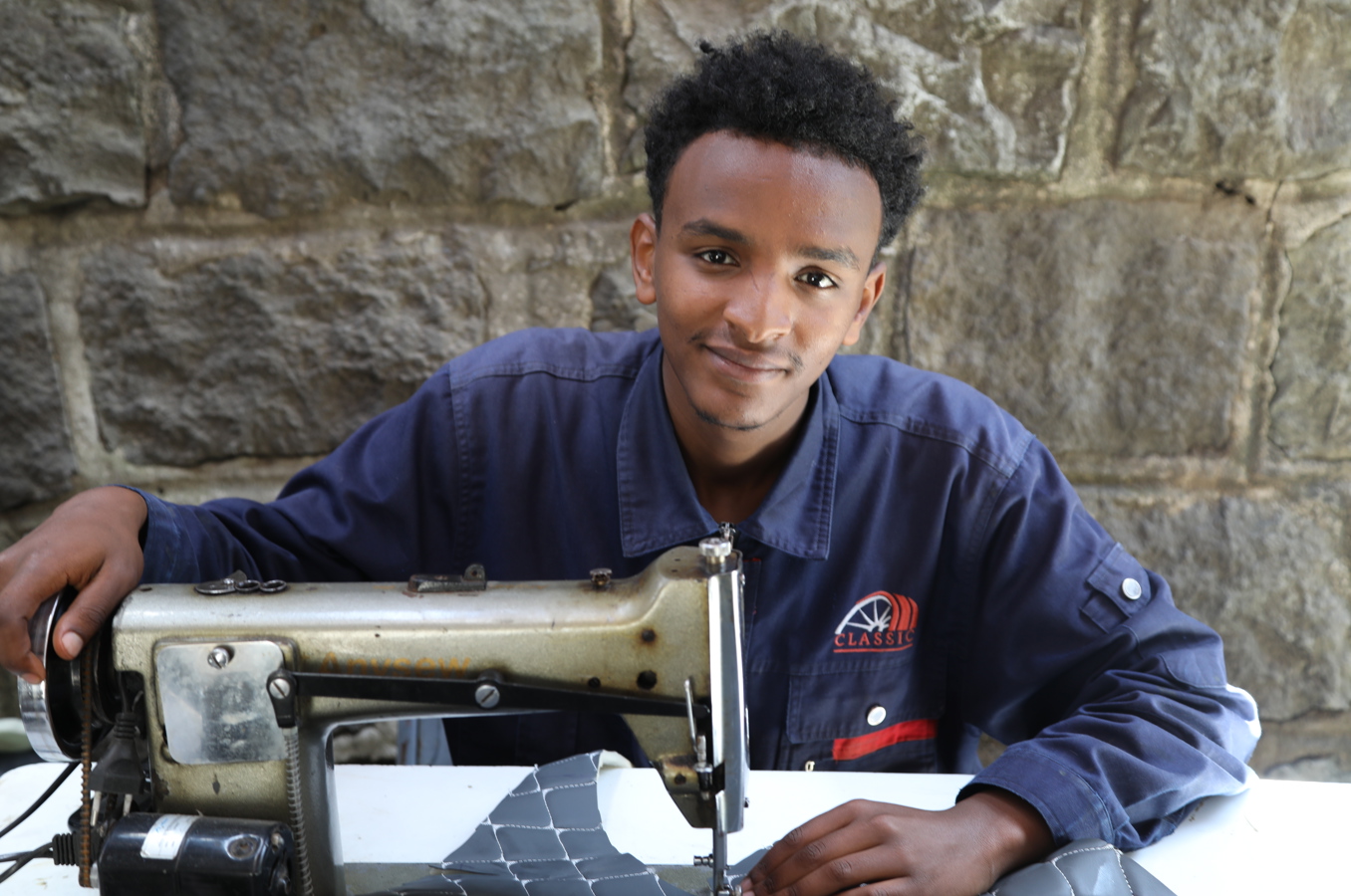 A young man in Ethiopia sits at a table with a sewing machine, smiling at the camera.