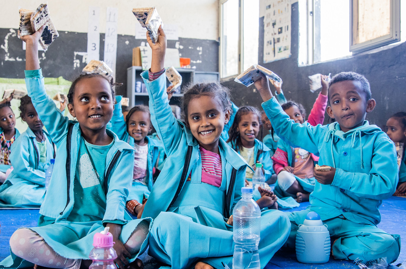 A group of children in an Ethiopian classroom sit on the floor and hold up their energy bars, smiling.