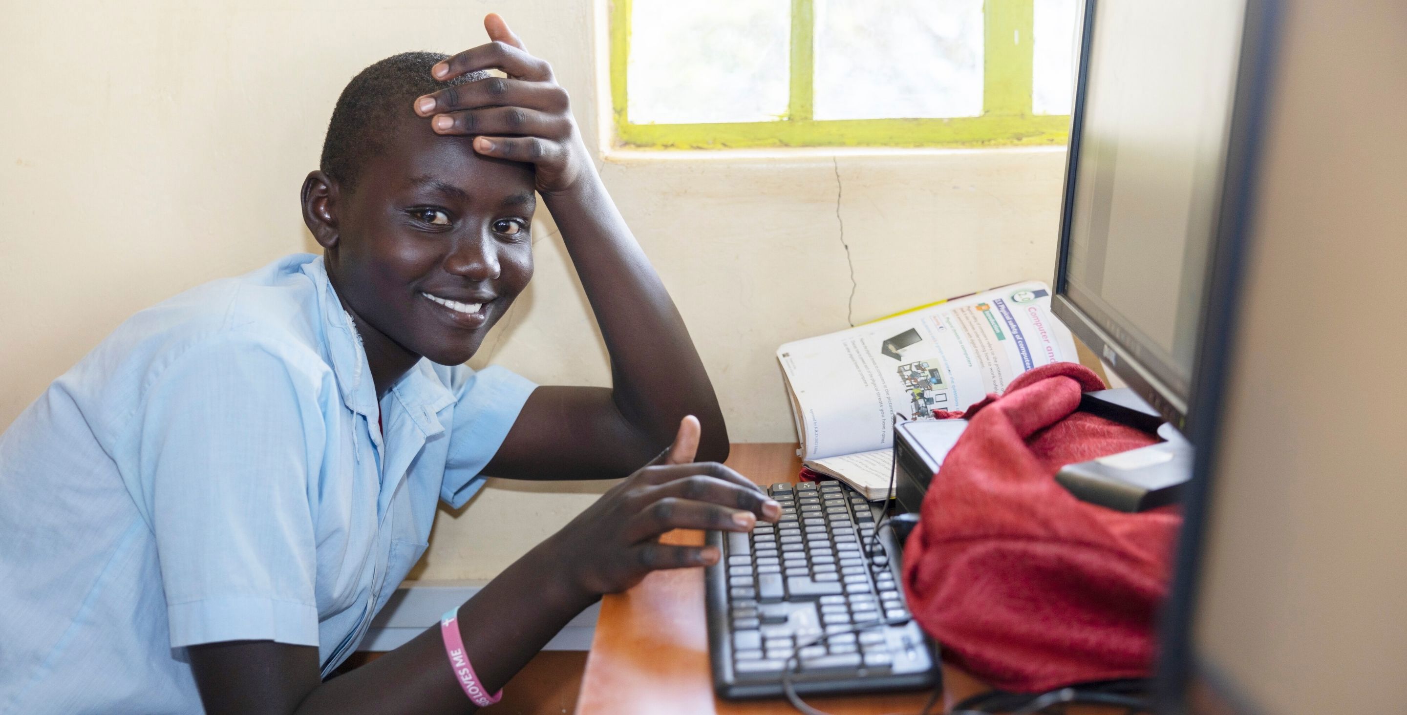 A young woman in Kenya sits at her desk in a computer lab, a book opened up at the computer beside her.