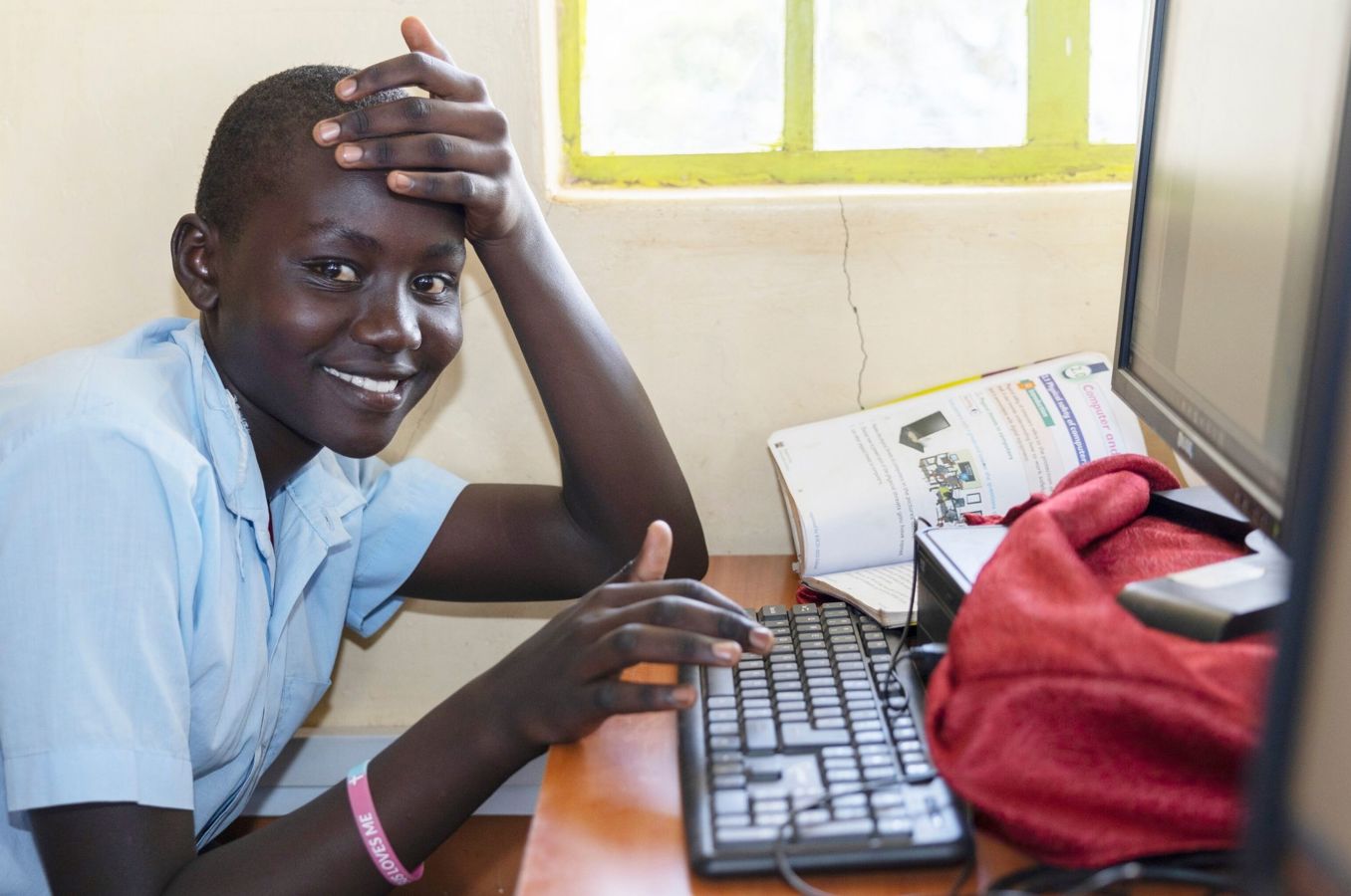 A young woman in Kenya sits at her desk in a computer lab, a book opened up at the computer beside her.