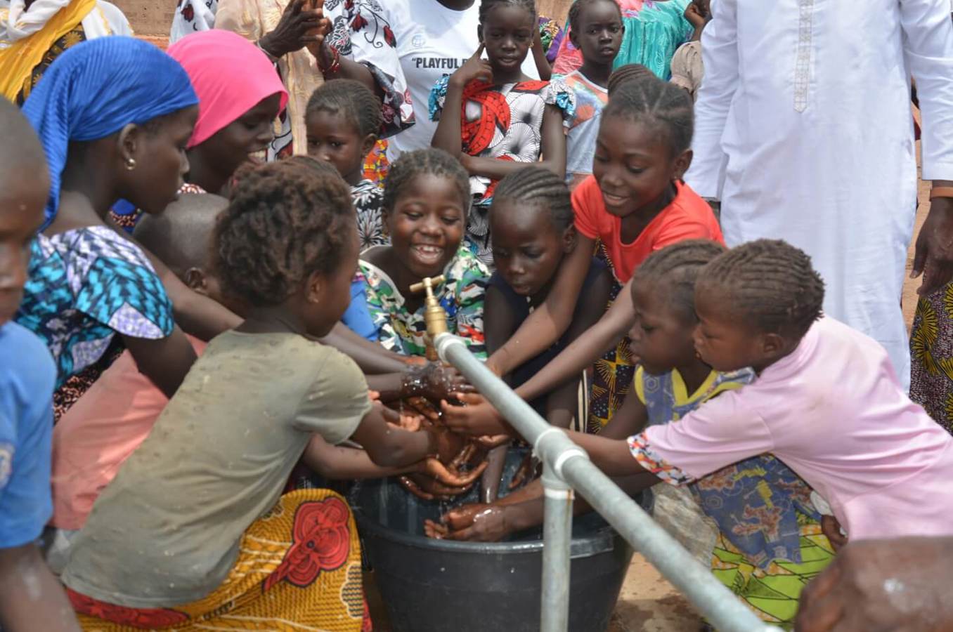 A group of children gather around a water faucet to wash their hands, smiling.