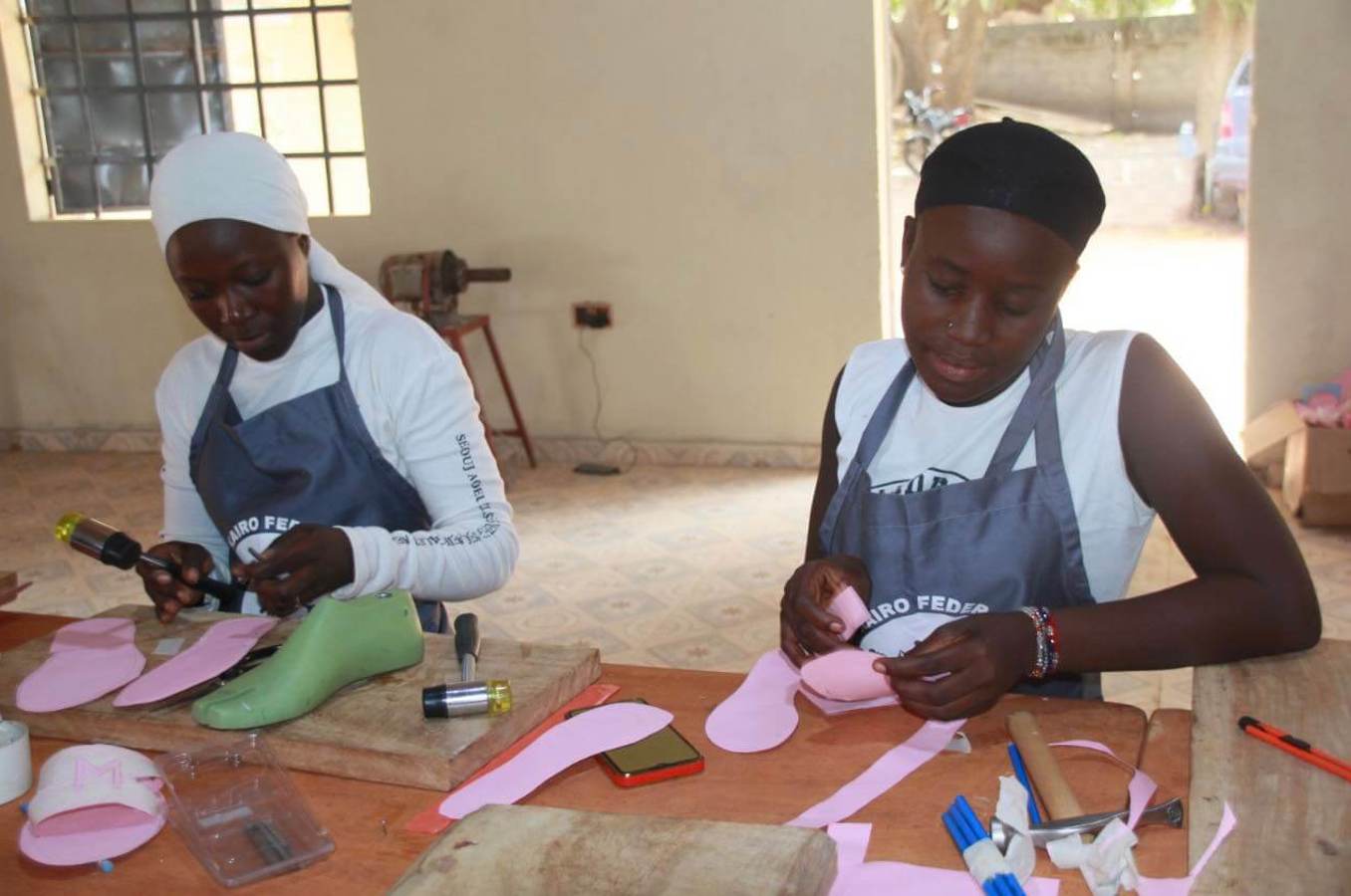 Two teenage girls sit at a table in The Gambia making shoes.