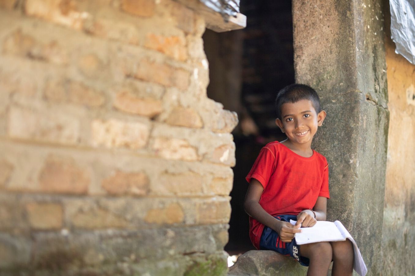A sponsored child in Sri Lanka sits in the doorway of his earthen home, writing a letter to his sponsor.