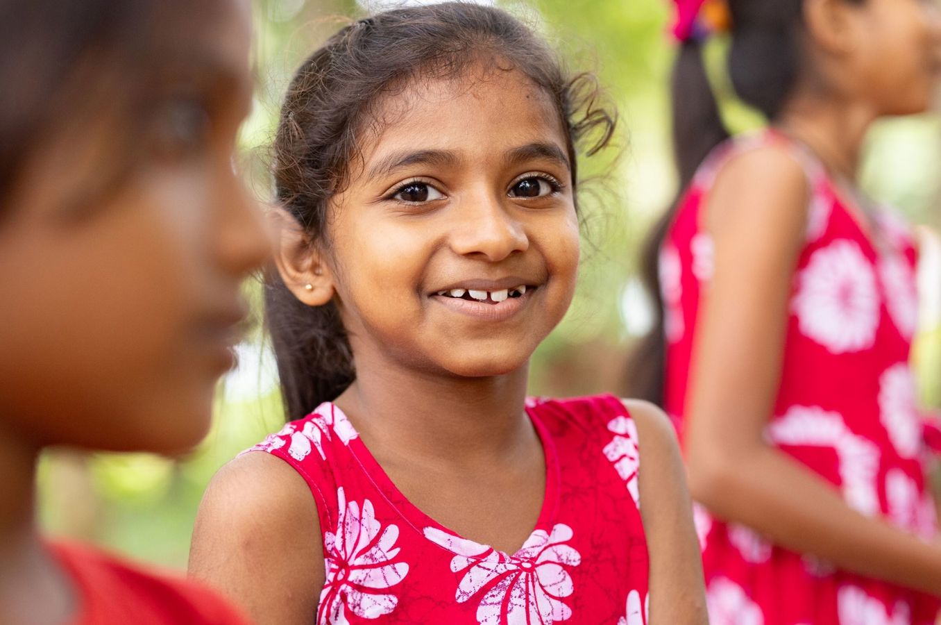 A girl wearing a red dress in Sri Lanka smiles outdoors while playing.